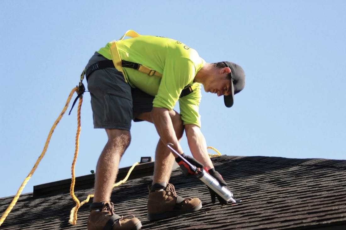 Roofer in a safety harness sealing shingles with a caulk gun